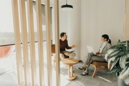 Two professionals engage in a lively discussion in a modern workspace filled with natural light. One is seated on a wooden chair, while the other works intently on a laptop.の写真素材