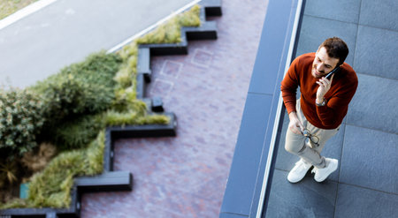 A young man stands on a sleek balcony while engaged in an animated phone conversation. Below him, a blend of contemporary design and lush greenery enhances the atmosphere.の写真素材