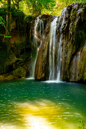 A breathtaking waterfall flows gracefully into a tranquil pool, surrounded by vibrant greenery and sun-dappled trees. Nature's beauty unfolds in a peaceful forest haven.の写真素材