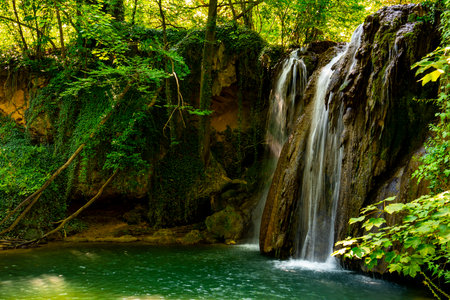A breathtaking waterfall flows gracefully into a tranquil pool, surrounded by vibrant greenery and sun-dappled trees. Nature's beauty unfolds in a peaceful forest haven.の写真素材