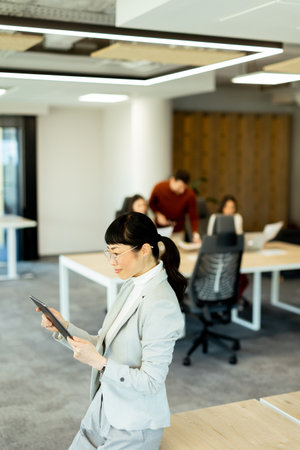 A focused Japanese woman in a suit reviews a document while colleagues discuss ideas in a modern office. The environment fosters creativity and teamwork in a vibrant workspace.の写真素材