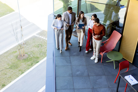 A diverse group of five professionals engage in lively discussions while reviewing digital tablets on a sleek terrace. Sunlight casts playful shadows on their attire.の写真素材