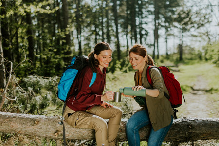 Two women take a moment to relax on a fallen log, sharing drinks and laughter in a vibrant forest full of life.の写真素材