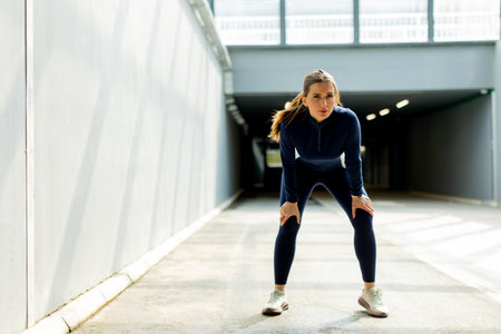 Focused and determined, a woman in athletic wear leans forward with hands on her knees, recovering in a sleek, sunlit hallway after exerting herself.の写真素材