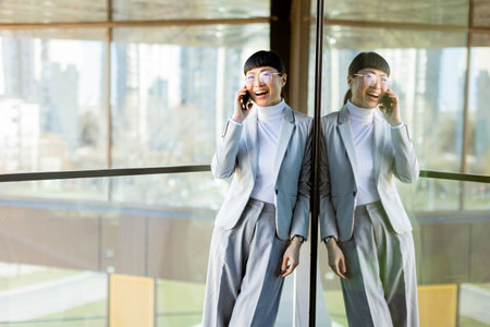 A fashionable Asian woman dressed in a tailored suit stands joyfully, chatting on her phone while leaning against a glass wall.の写真素材