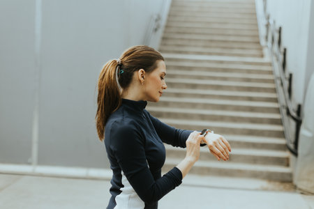 Focused individual monitors her smartwatch while preparing for an energetic morning run near a stylish staircase in an urban environment.の写真素材