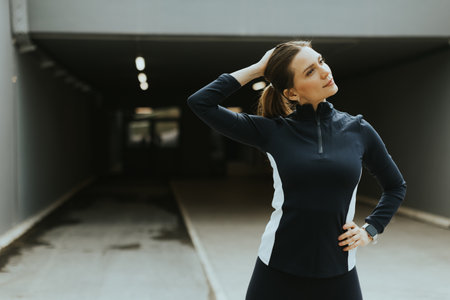 A young woman poses confidently with one hand on her neck while standing in a modern urban setting, ready for her workout session.の写真素材