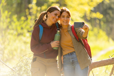 Two friends take a cheerful selfie amidst the blooming nature, surrounded by lush trees and warm...