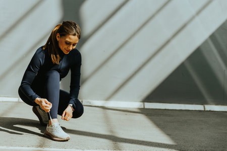 A woman crouches on a smooth surface, adjusting her shoelaces with sunlight streaming through shadows, preparing for an active day.の写真素材