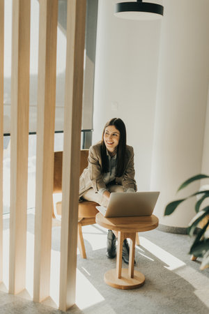 In a contemporary office, a focused businesswoman types on her laptop while seated on a chic chair, surrounded by natural light filtering through the windows, enhancing her concentration.の写真素材