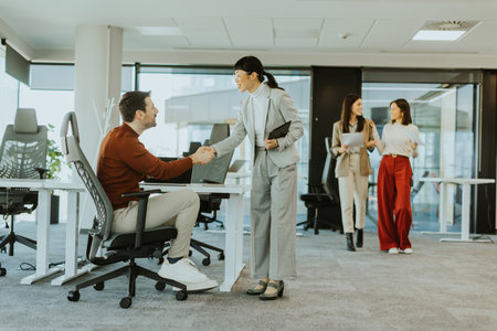 Bright office environment sees a joyful handshake between a male and female professional. They...