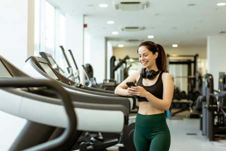 In a modern gym filled with sunlight, a young woman smiles as she checks her phone, taking a break...