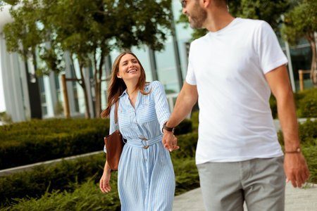 A couple walks hand in hand with smiles on their faces, surrounded by lush greenery in a lively urban park on a bright day.の写真素材