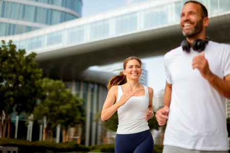 Two joggers share smiles as they run through a vibrant urban park, surrounded by sleek buildings and lush landscaping on a sunny morning.の写真素材