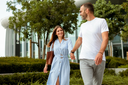 A man and a woman walk hand in hand through lush greenery, sharing smiles and laughter in a vibrant city environment under a clear sky.の写真素材