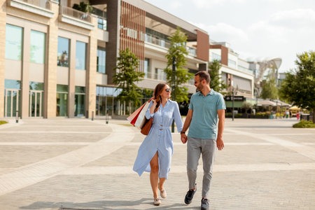 Two people stroll hand-in-hand through a vibrant shopping area, carrying bags and enjoying the warm sunshine on a delightful afternoon.の写真素材