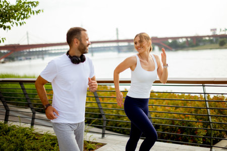 A couple enjoys jogging side by side along a scenic river pathway, exchanging smiles and encouraging words amidst the sunlight.の写真素材