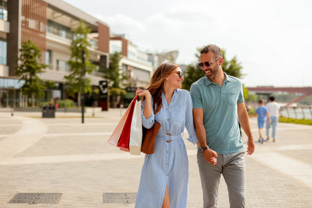 On a bright day, a couple walks hand in hand along a charming riverside path, carrying shopping bags filled with goodies, smiling and laughing.の写真素材