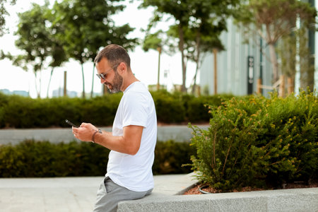 Bright sunshine illuminates a casual man dressed in a white t-shirt and sunglasses, absorbed in his smartphone while resting in a tranquil park.の写真素材