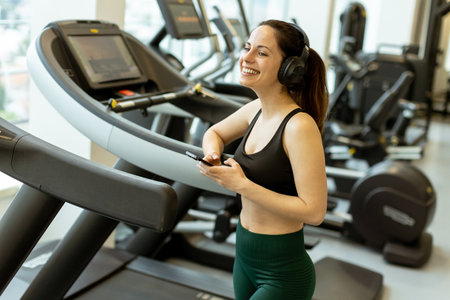 A woman in workout attire stands on a treadmill, smiling as she holds a phone, clearly enjoying her...