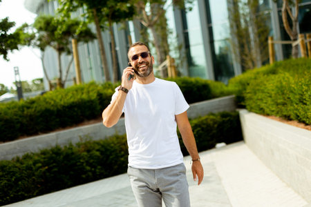 Sunlight fills the urban park as a cheerful man in a white shirt smiles and chats on his phone, surrounded by greenery and modern buildings.の写真素材
