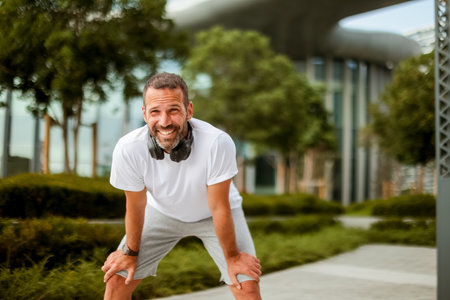 A cheerful man leans forward, catching his breath in a trendy green space, showcasing relaxation and joy after exercising outdoors.の写真素材