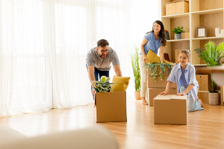 Three friends enthusiastically arrange their new living room, unpacking boxes filled with greenery and cozy decorations, embraced by natural light.の写真素材