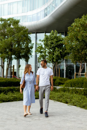 A man and woman walk hand in hand through a lush urban park, surrounded by greenery and contemporary buildings, enjoying their time together.の写真素材