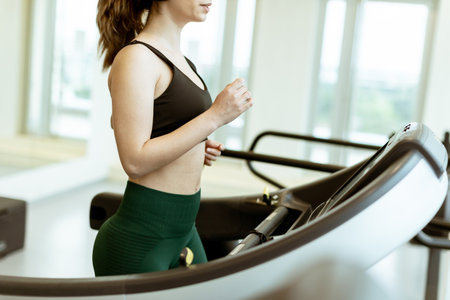 A woman enjoys her workout, focused on the treadmill, earbuds in place, energizing herself in a...