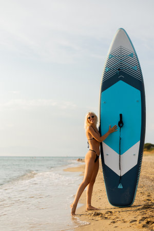 A woman with sunglasses stands on the sandy shore, leaning against a bright paddleboard. The serene water reflects the warm colors of the sunset as waves gently lap at the beach.の写真素材