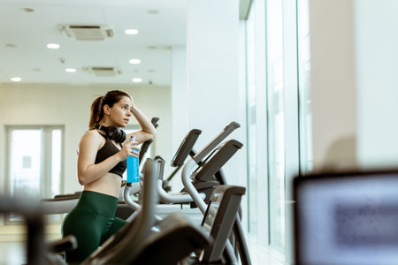 In a sleek, bright gym, a woman pauses her intense workout on an elliptical machine to hydrate,...