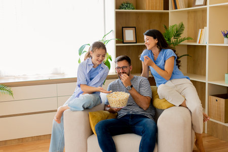 A joyful family shares a cozy evening on the couch, munching popcorn and laughing together. The warm...