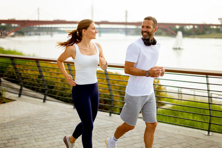 Two friends jog together beside the river, sharing smiles and laughter as they enjoy the scenic view during their morning exercise routine on a sunny dayの写真素材