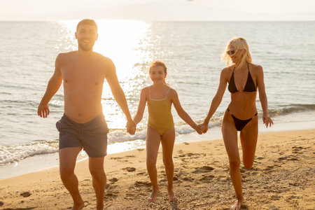 A family of three plays on the sandy shore, hand in hand, enjoying the warm sun and gentle waves under a serene sky.の写真素材