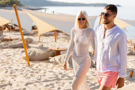 A cheerful couple walks together on the sandy beach, dressed in light summer attire, surrounded by relaxing sunbeds and a peaceful atmosphere.の写真素材