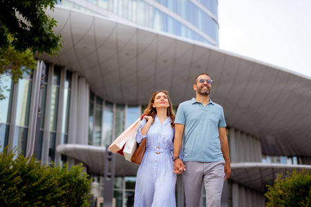 A young couple strolls hand in hand, smiling and enjoying the sunny weather, surrounded by modern architecture and holding shopping bags, creating a cheerful atmosphere of leisure.の写真素材