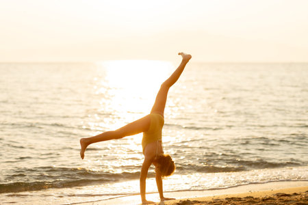 A cheerful young person performs a handstand on the sandy beach as the sun sets over the calm ocean...