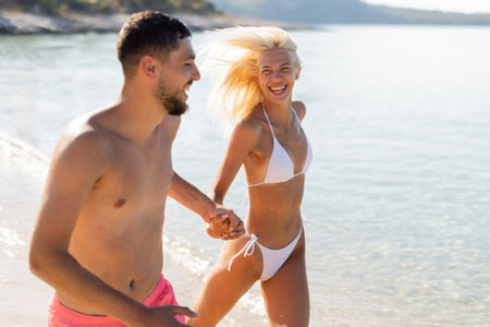 A couple enjoys a playful moment at the beach, holding hands and laughing as they run through shallow waves of crystal clear water.の写真素材