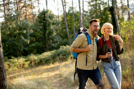 Two friends walk along a forest trail on a sunny afternoon, carrying backpacks and smiling. The vibrant greenery creates a lively atmosphere as they enjoy each other's company.の写真素材
