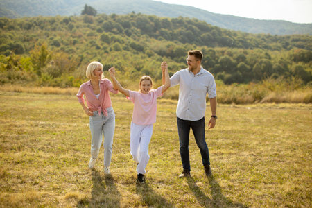 A joyful family strolls through a sunny field, holding hands and smiling. The scene features lush green hills and a clear sky, creating a warm afternoon atmosphere.の写真素材
