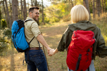 Two people hike together on a sunny day in a forest, holding hands and smiling. Their backpacks show they are ready for a day of adventure and exploration in nature.の写真素材