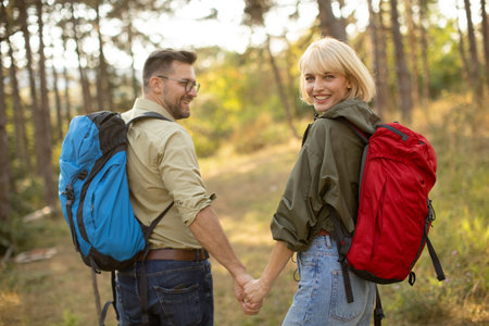 Two people hike together on a sunny day in a forest, holding hands and smiling. Their backpacks show they are ready for a day of adventure and exploration in nature.の写真素材