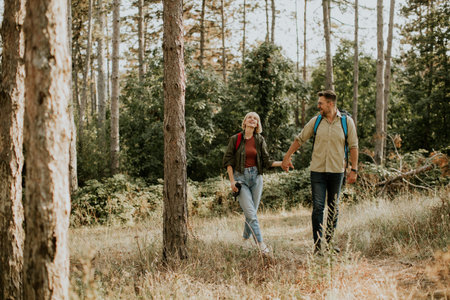 A couple walks hand in hand along a forest trail, surrounded by tall trees and lush greenery. The sun filters through foliage, creating a warm and inviting atmosphere as they explore nature.の写真素材