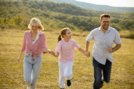 A family of three happily runs hand in hand through a sunny field. They are enjoying a carefree...