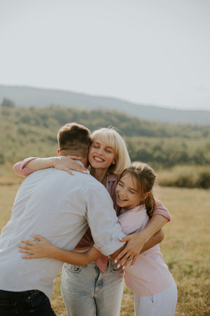 A joyful family shares a warm embrace in a green countryside field under a sunny sky. Smiles and laughter highlight this precious moment of togetherness.の写真素材