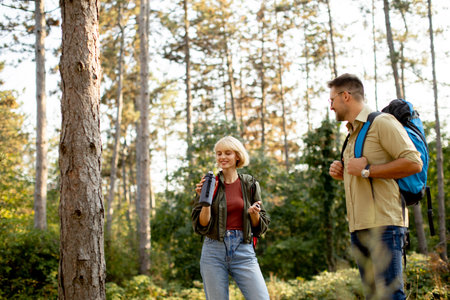 Two young adults stand in a lush forest, engaging in conversation as they take a break from hiking. The sun filters through the tall trees, creating a serene atmosphere.の写真素材