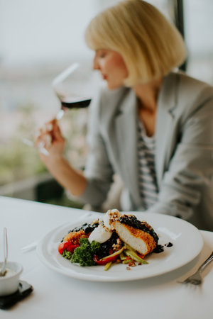 A woman in a gray blazer sips red wine while dining at an upscale restaurant. A plate of gourmet food is in front of her, and the city skyline is visible through the window.の写真素材