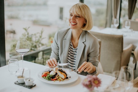 A smiling woman sits at a table in a chic restaurant, savoring a beautifully presented dish. The setting features large windows, allowing bright sunlight to flood the space.の写真素材