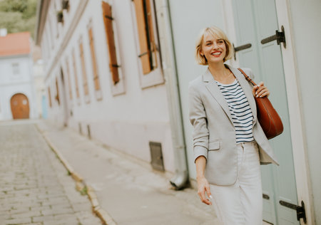 A confident woman walks along a narrow cobblestone street surrounded by beautiful historic buildings. She wears a light blazer and striped shirt, enjoying the sunny weather.の写真素材