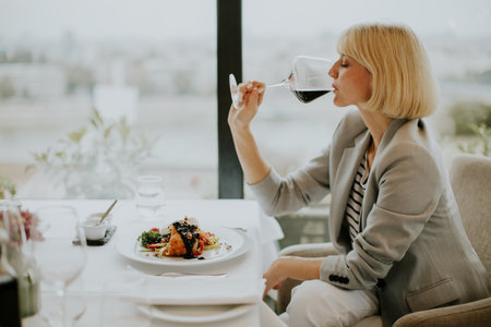 A woman in a gray blazer sips red wine while dining at an upscale restaurant. A plate of gourmet food is in front of her, and the city skyline is visible through the window.の写真素材